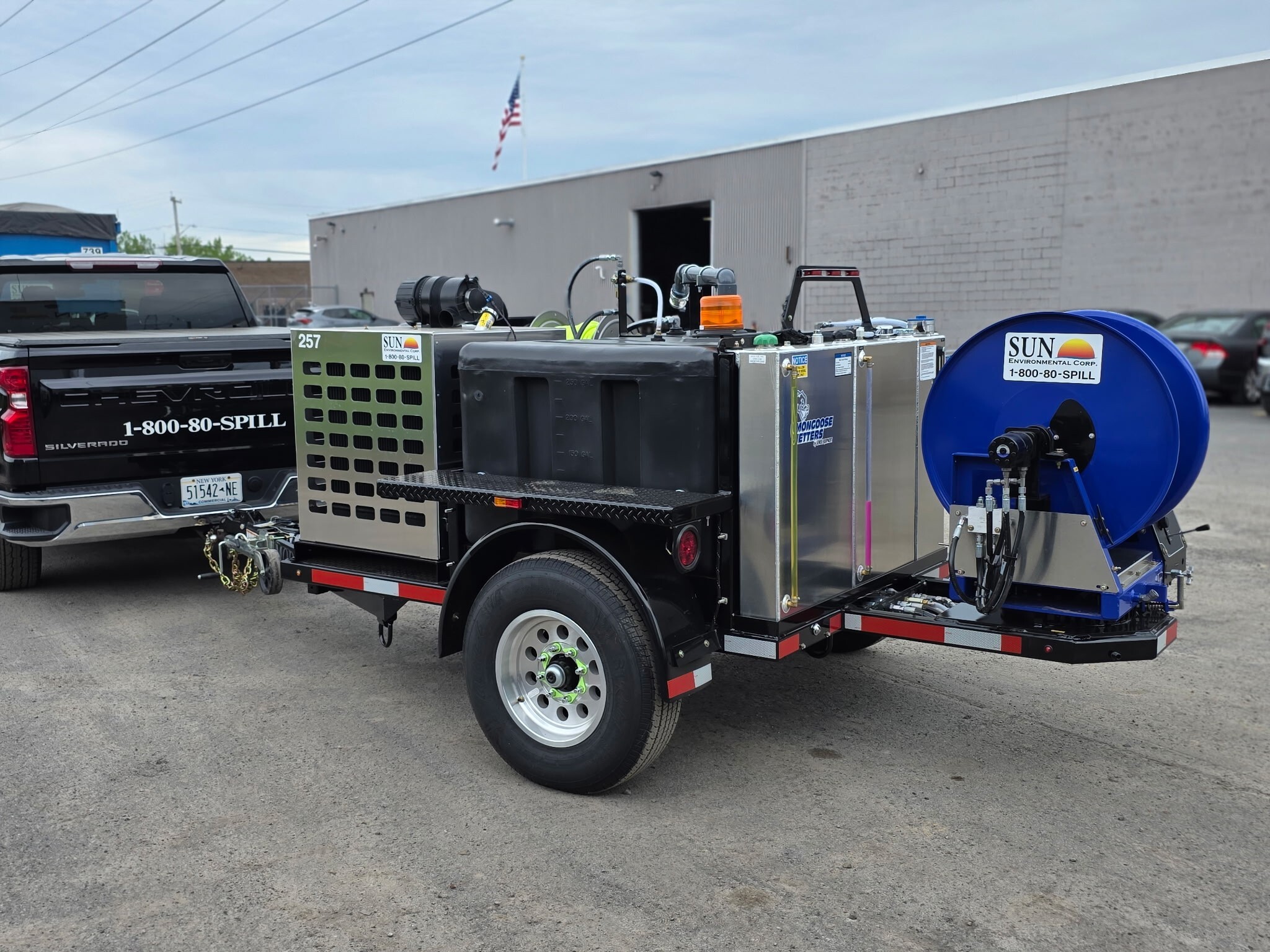 A white semi-truck with the Sun Environmental Corp logo on the door uses a hydraulic system to lower a bright blue roll-off bin to the ground